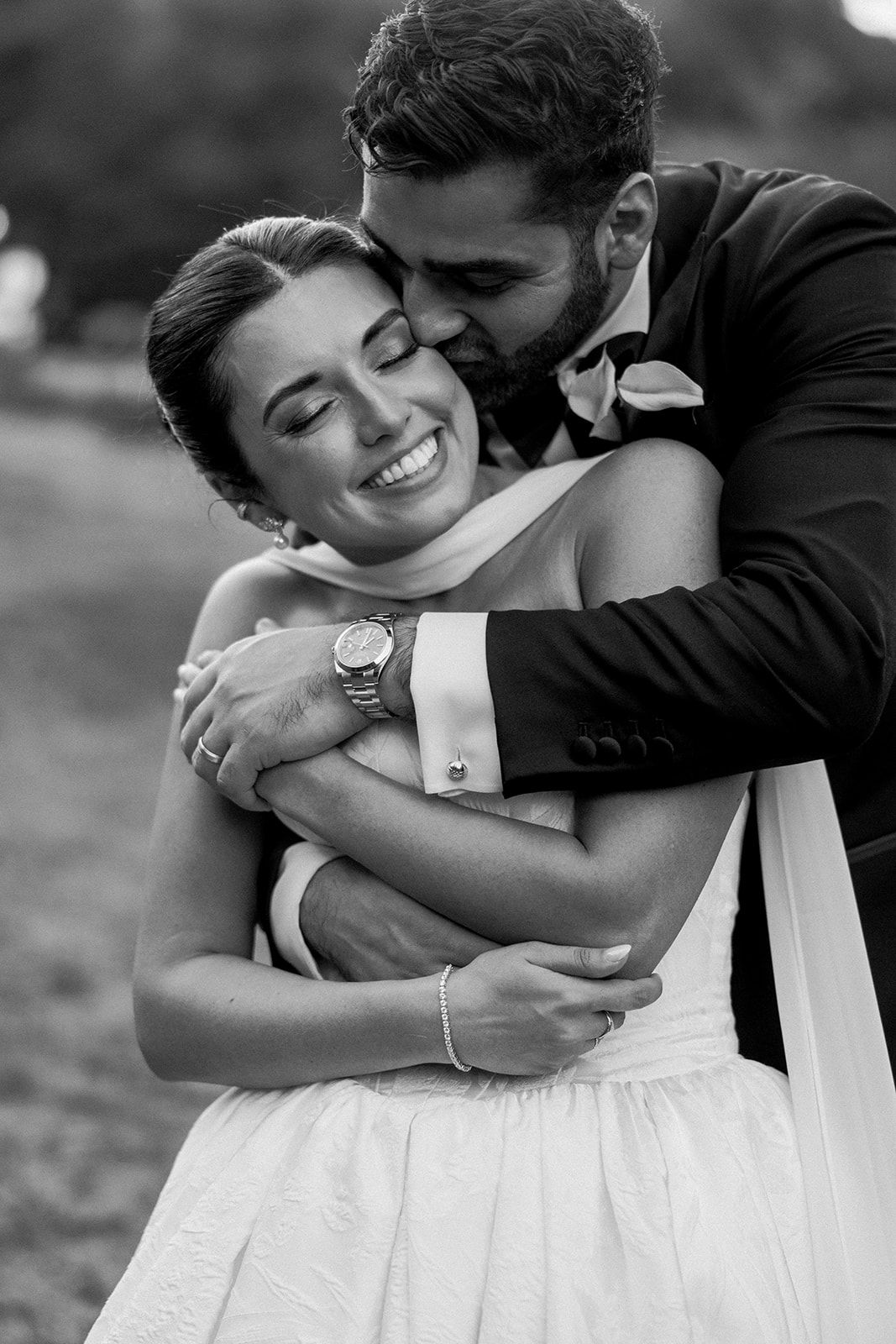 A tender black and white wedding photograph showing an affectionate embrace between newlyweds outdoors.