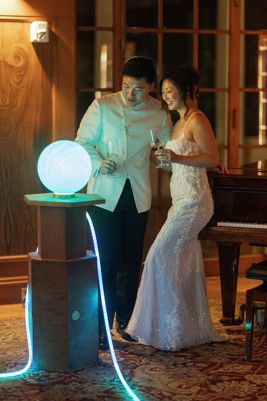 A bride and groom in wedding attire smile while holding champagne glasses near a glowing orb display at their reception.