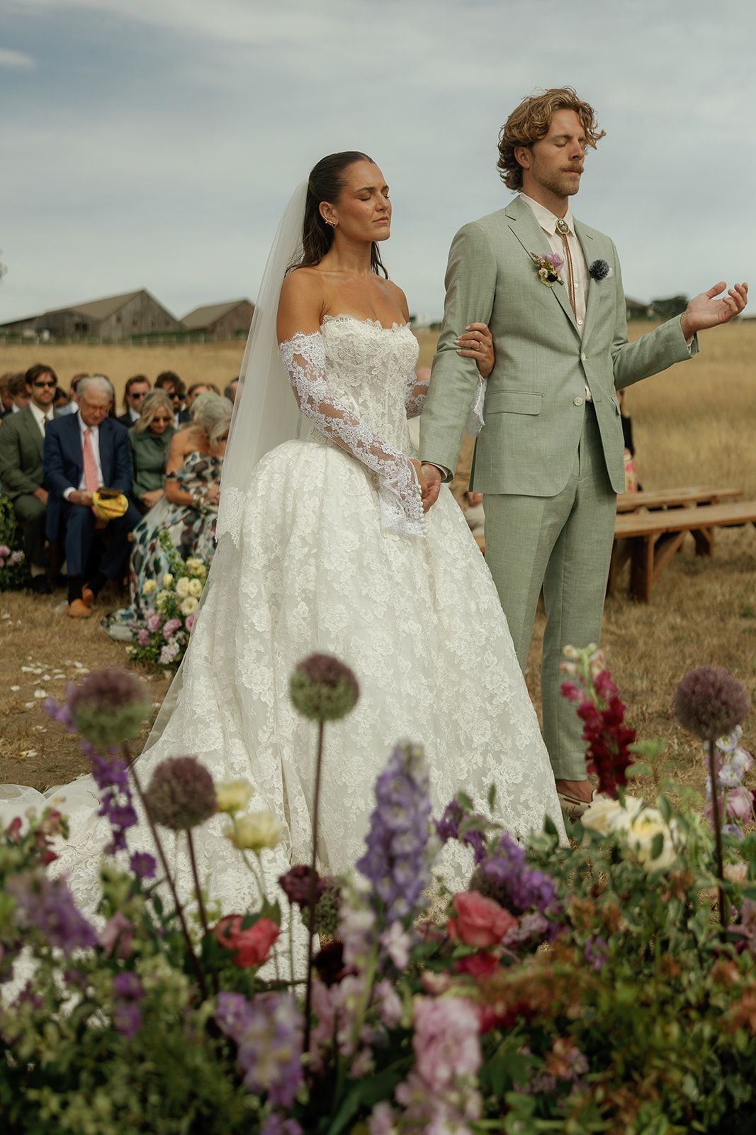 Bride in lace ball gown and groom in sage suit walk down outdoor aisle surrounded by wildflowers at rustic countryside wedding.
