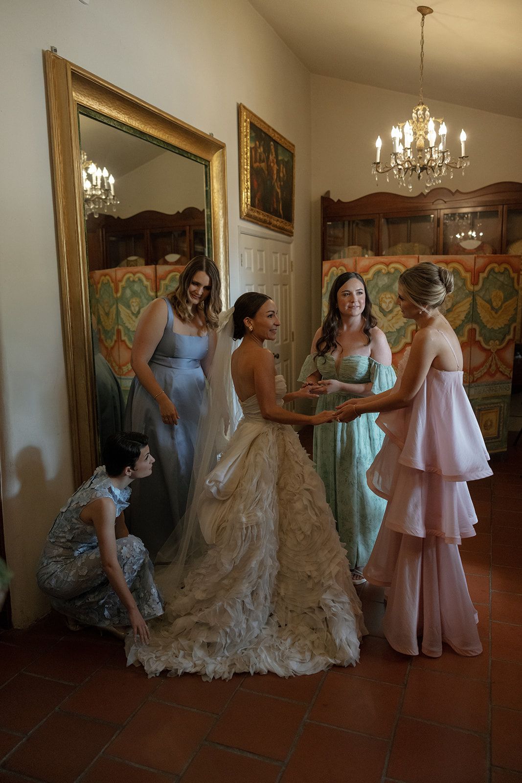 Wedding preparations in an elegant room with bridesmaids and gowns in pink and blue helping with dress fittings.