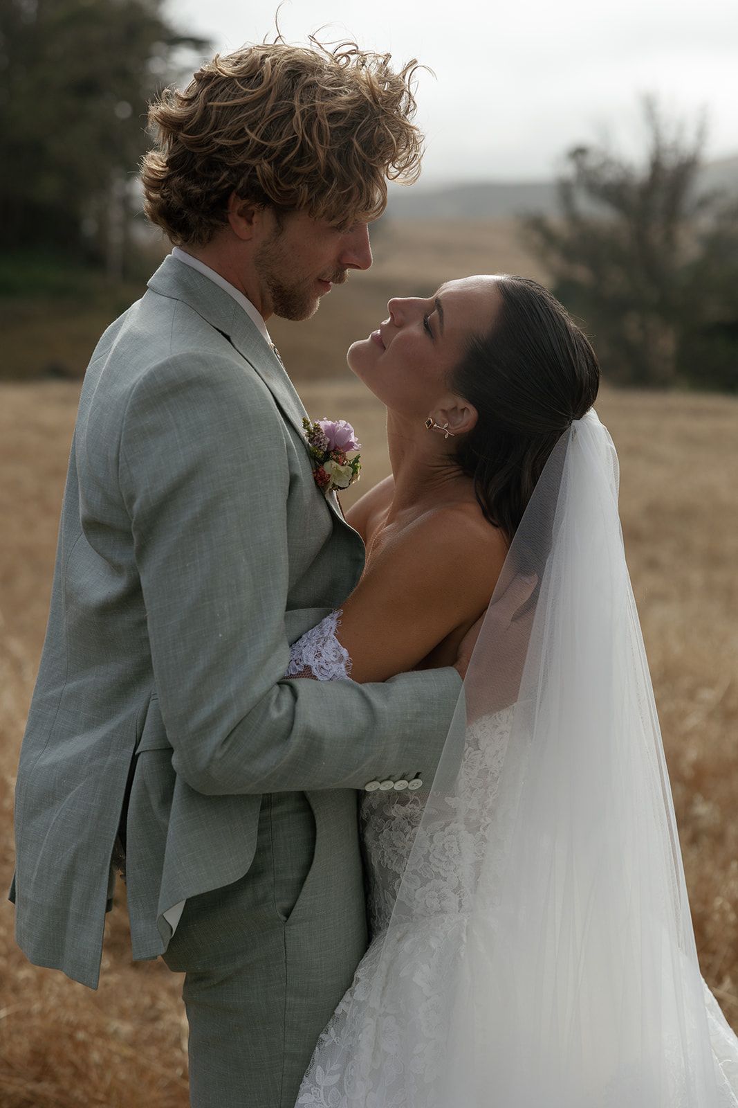 A bride and groom share a romantic embrace in a golden field, with the bride in a lace gown and veil.