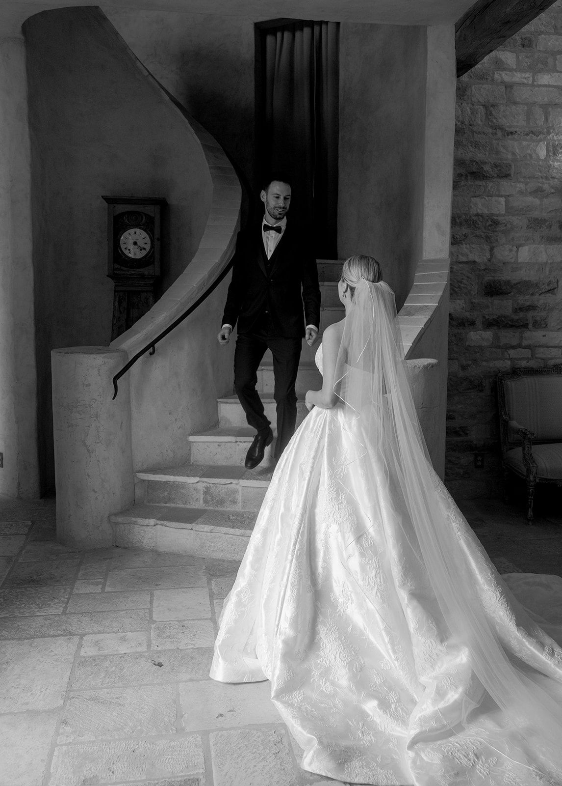 Black and white wedding photo of bride in ball gown facing groom descending elegant spiral staircase.