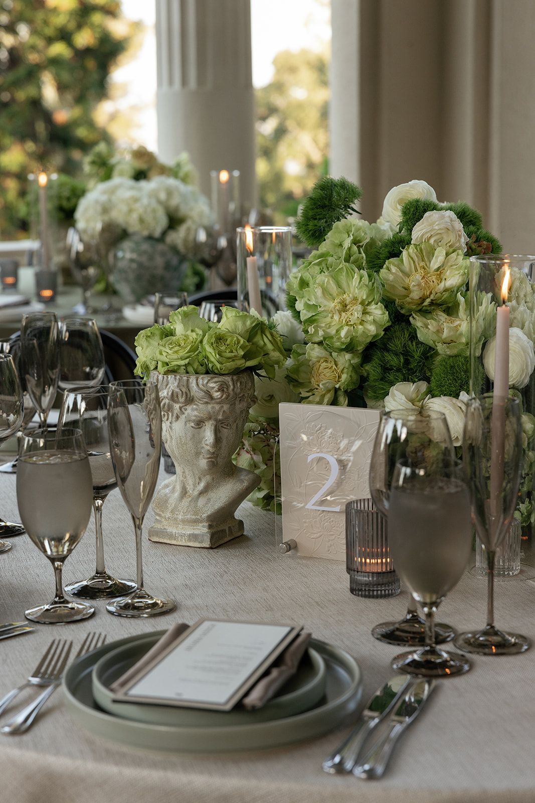 Elegant wedding table setting with white floral centerpieces, silver place settings, and table numbers on a neutral tablecloth.