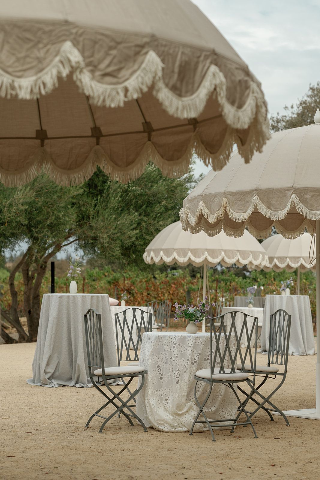 Elegant outdoor dining setup with fringed parasols, metal chairs, and lace-draped tables in a vineyard setting.