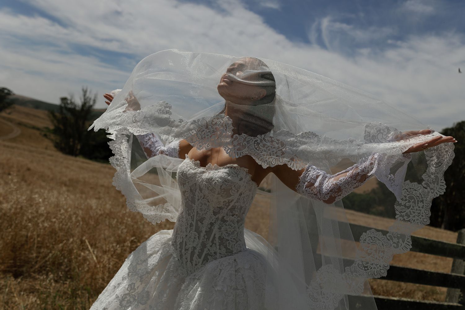 Bride in white lace wedding dress with veil blowing in wind, standing in golden field under cloudy sky.