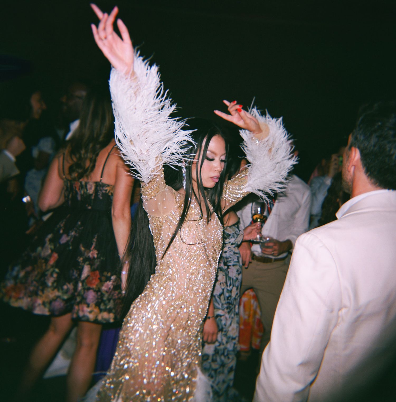 Person in a sparkly beige dress dances with feather accessories at a crowded nightclub party.