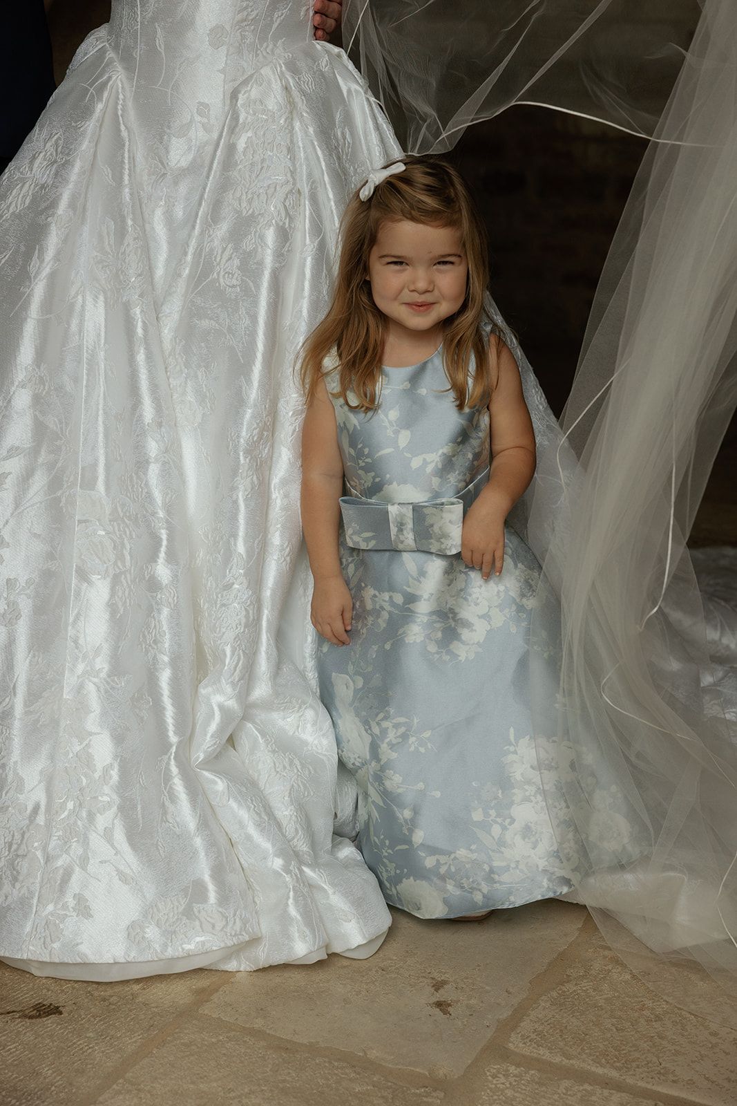 A young flower girl in a light blue floral dress smiles beside a bride in a white wedding gown.
