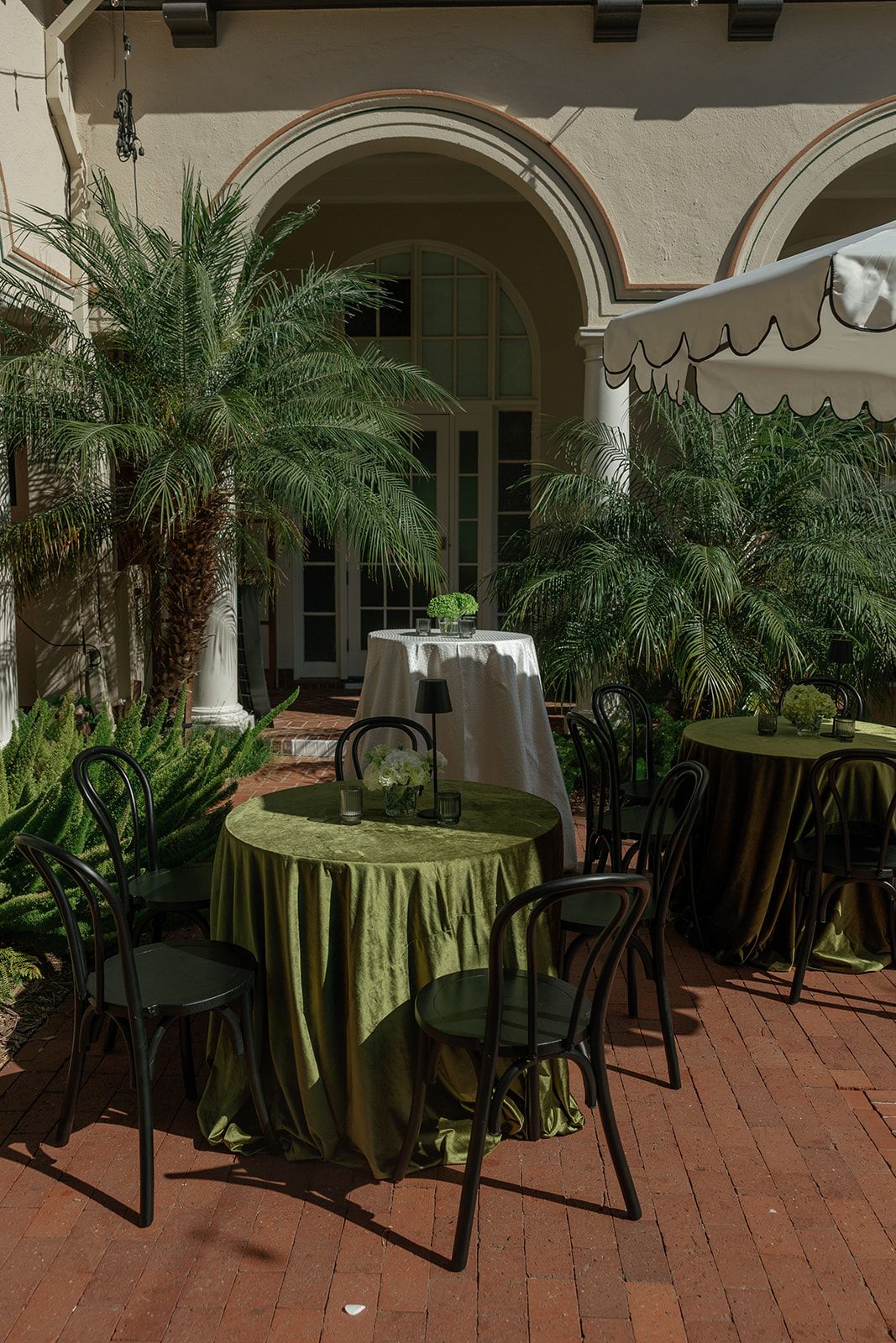 Elegant outdoor dining area with green tablecloths and palm trees under arched columns on brick patio.