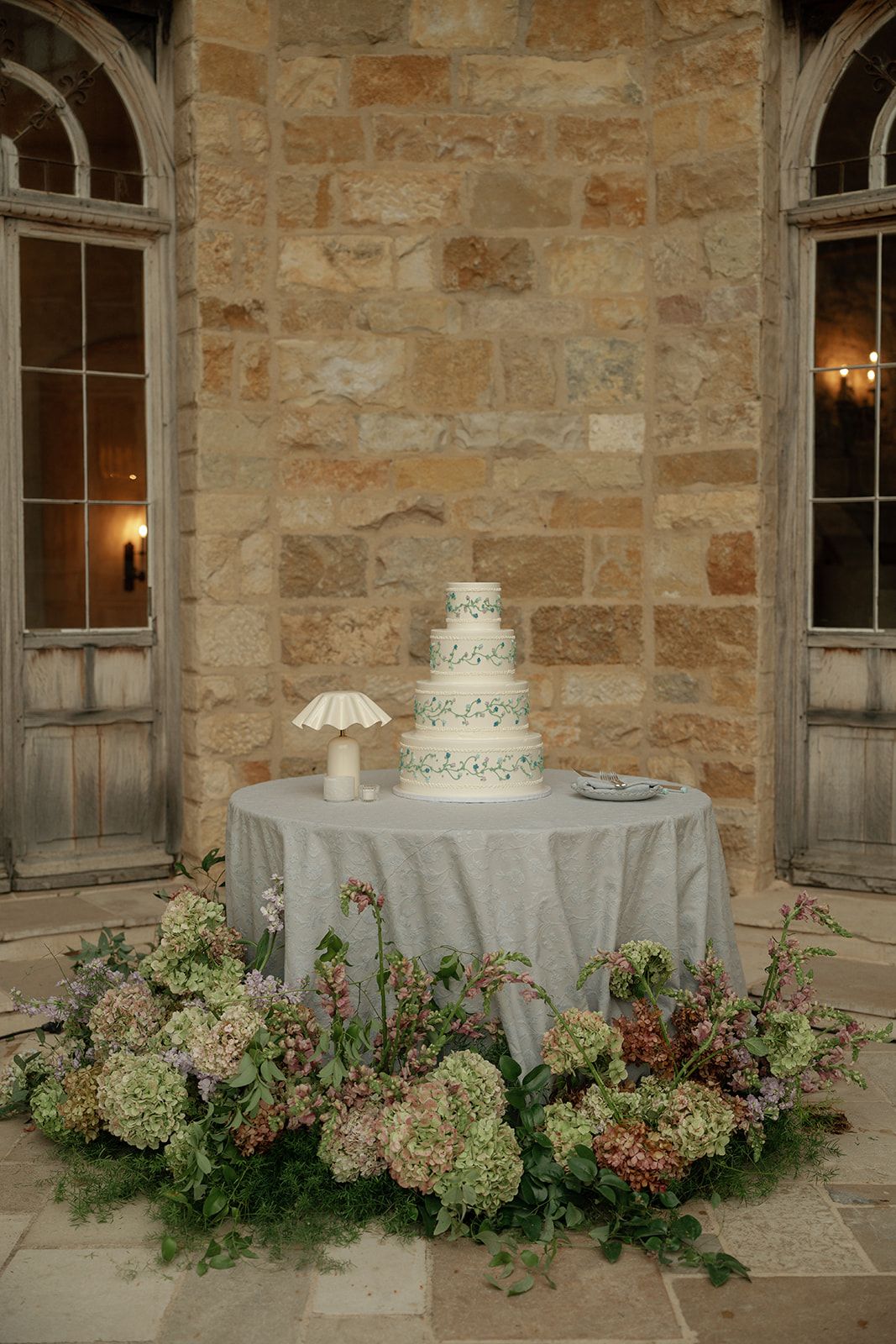 Elegant four-tier wedding cake on draped table surrounded by lush hydrangea florals against stone wall.