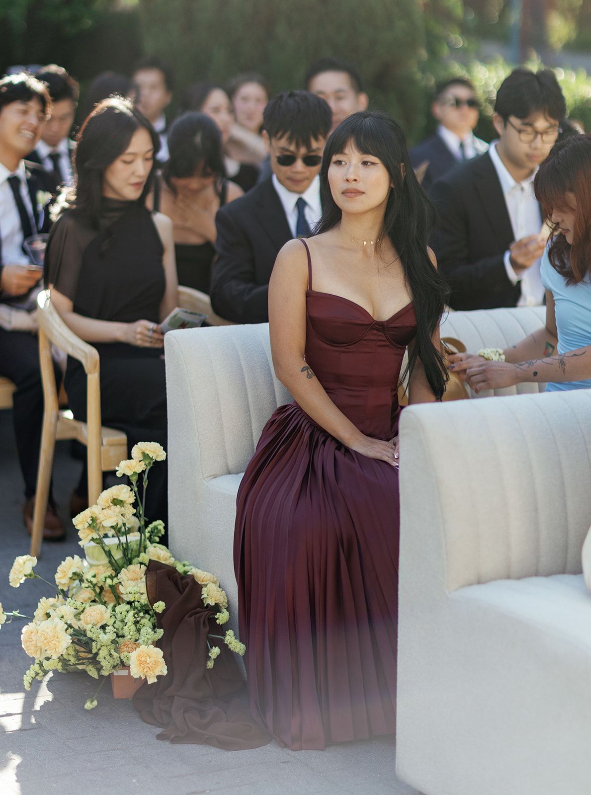 Woman in burgundy pleated gown seated at outdoor wedding ceremony with floral arrangement nearby.
