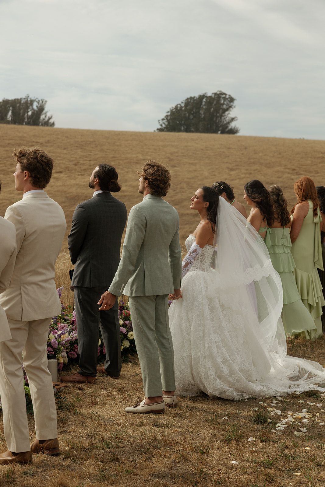 Bride in white gown and groom in sage suit stand with wedding party outdoors in a golden field ceremony.
