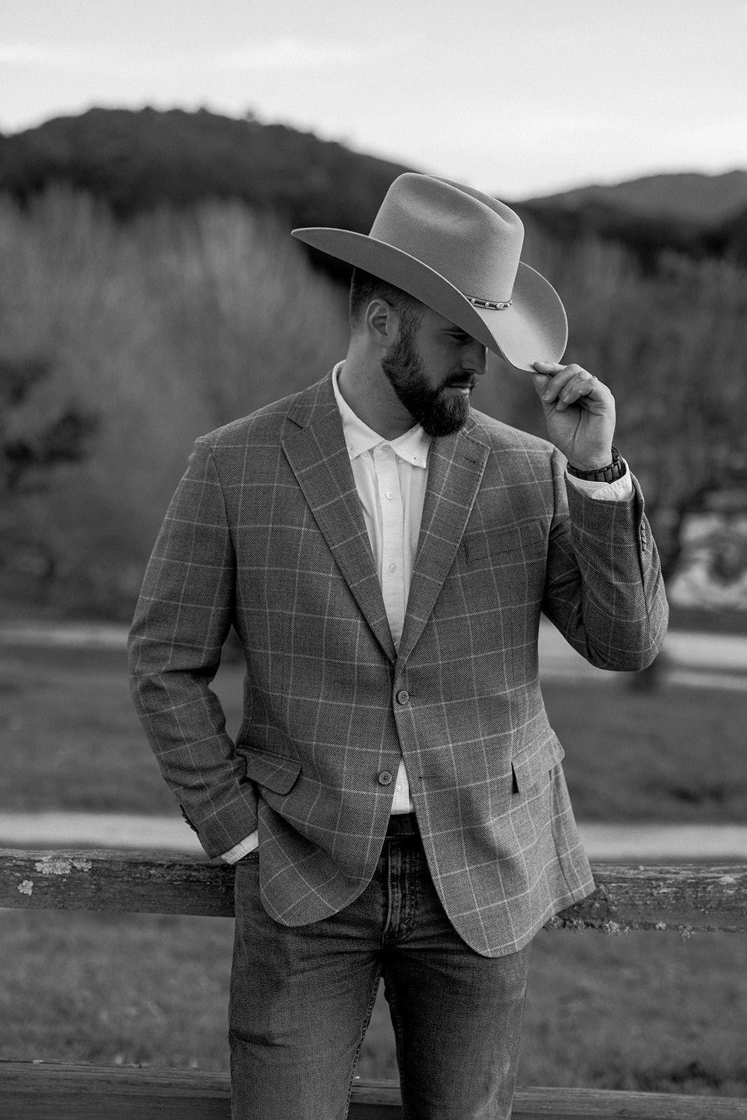 Black and white photo of someone wearing a plaid blazer and cowboy hat posing outdoors in a rustic setting.