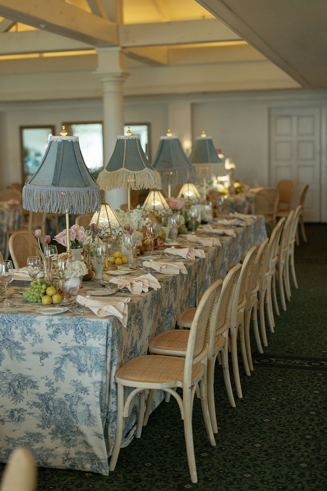 Elegant dining table set with blue and white toile tablecloth and classic lampshades for a formal event setup.