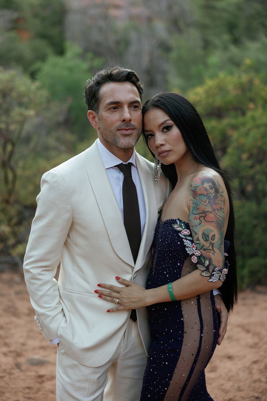 A couple poses together outdoors wearing formal attire, with one in a white suit and another in a sparkly navy dress.