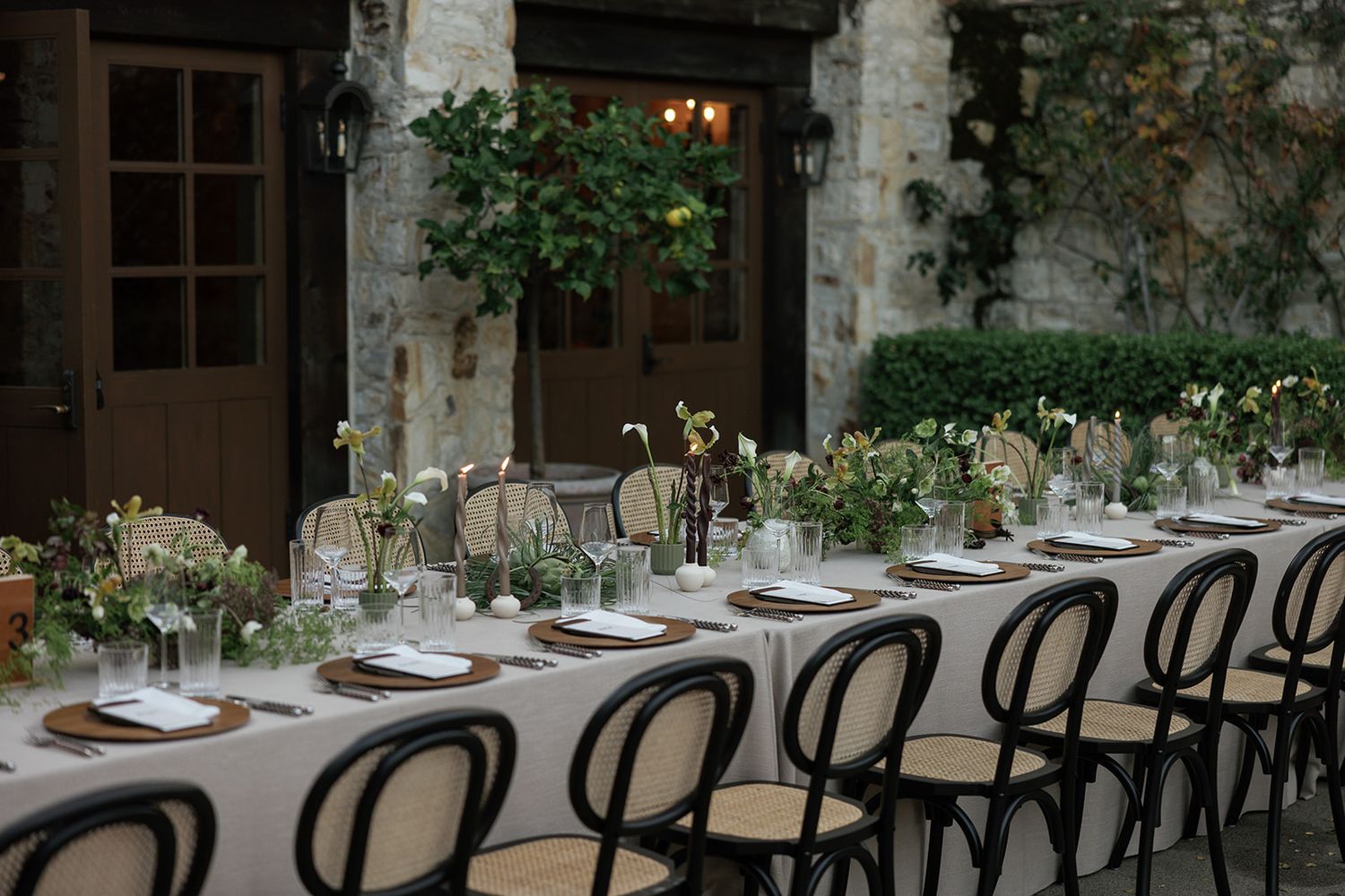 A rustic outdoor dining setup with long wooden tables and black chairs arranged under stone archways and climbing vines.