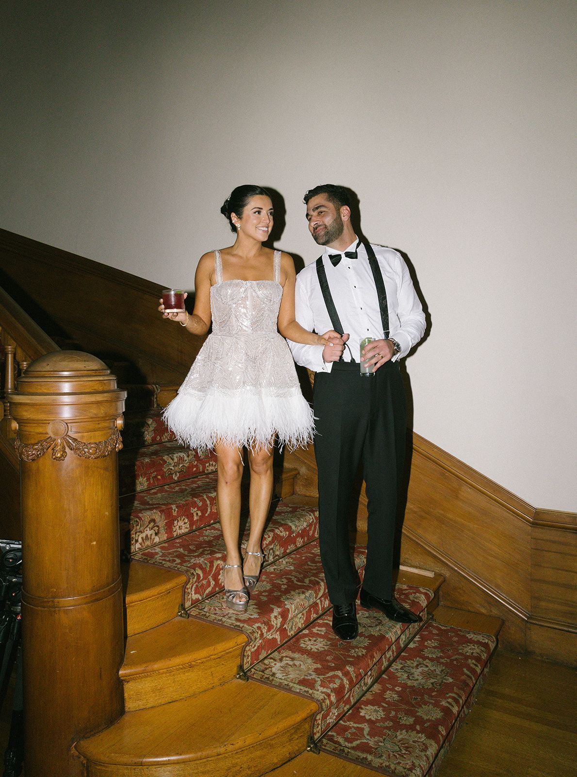 Formal couple stands on vintage staircase with ornate carpet runner and wooden newel post during special occasion.