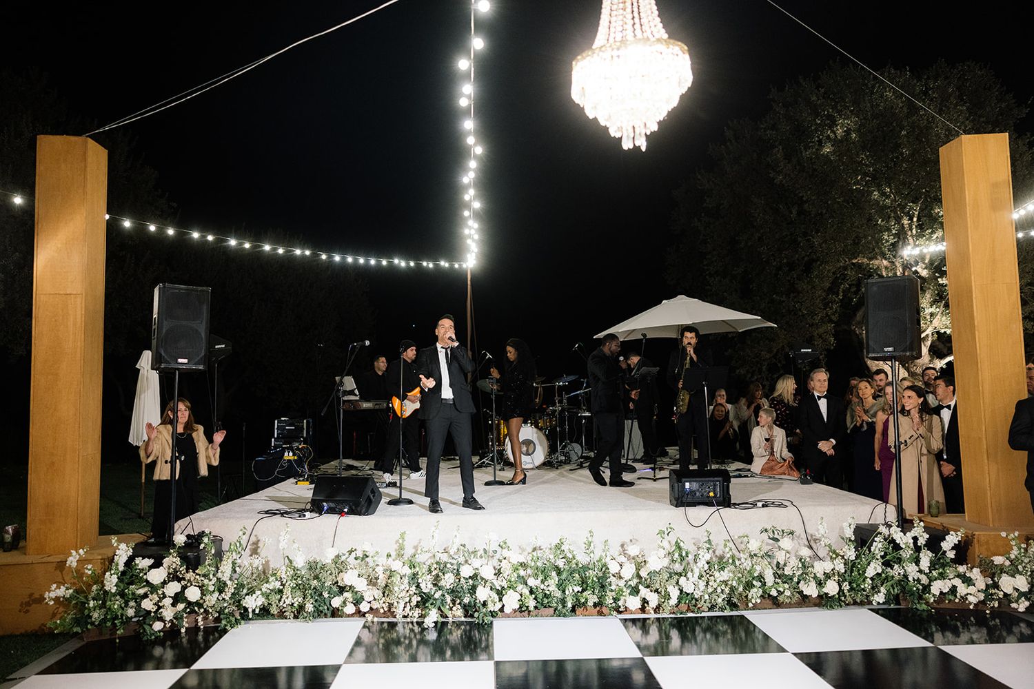 An outdoor wedding reception at night with string lights, a crystal chandelier, and white floral arrangements on the dance floor.