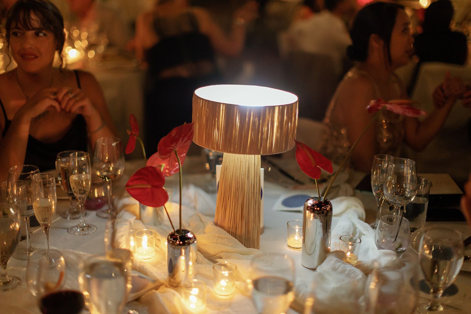 An elegant dining table setting with a wood and metal table lamp glowing warmly among wine glasses and candlelight.
