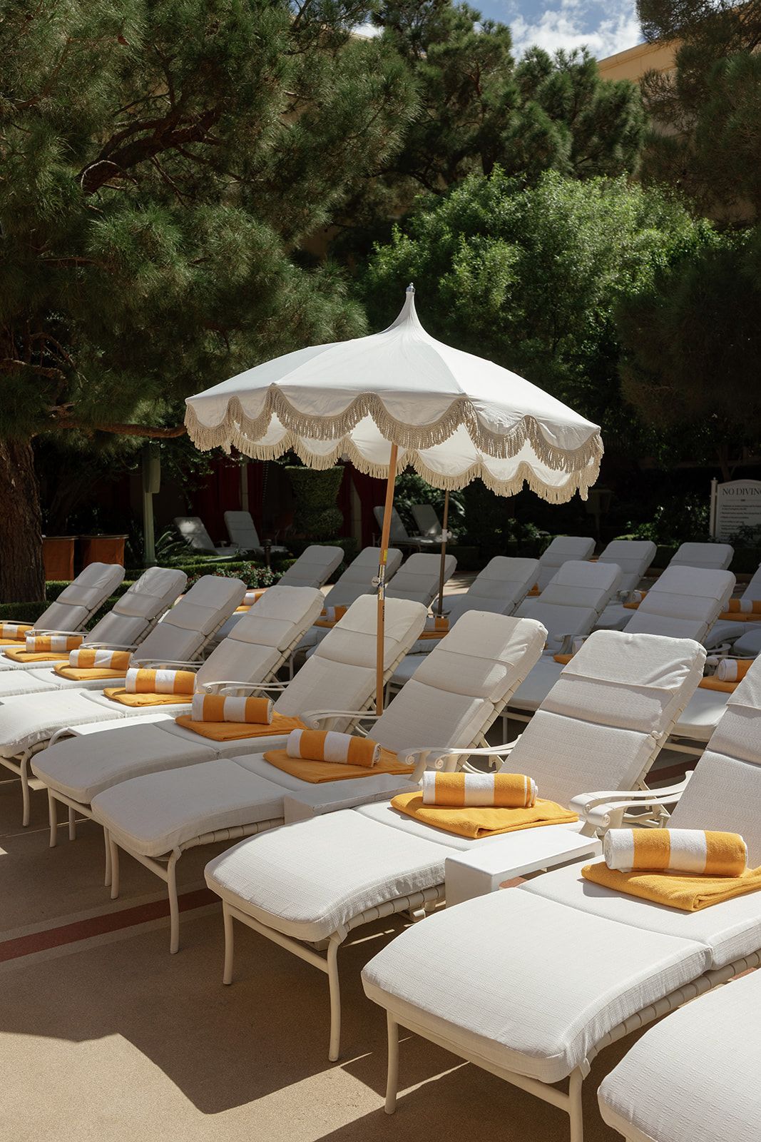 Luxury poolside lounge chairs with white cushions, yellow towels, and a fringed umbrella surrounded by lush green trees.