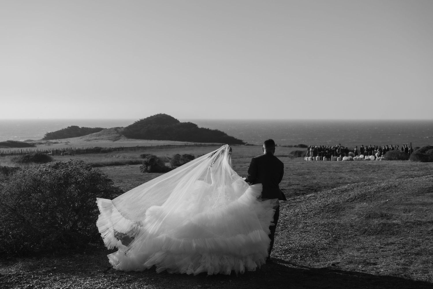 Black and white photo of a bride in a flowing gown with a long veil on a coastal hilltop with ocean views.