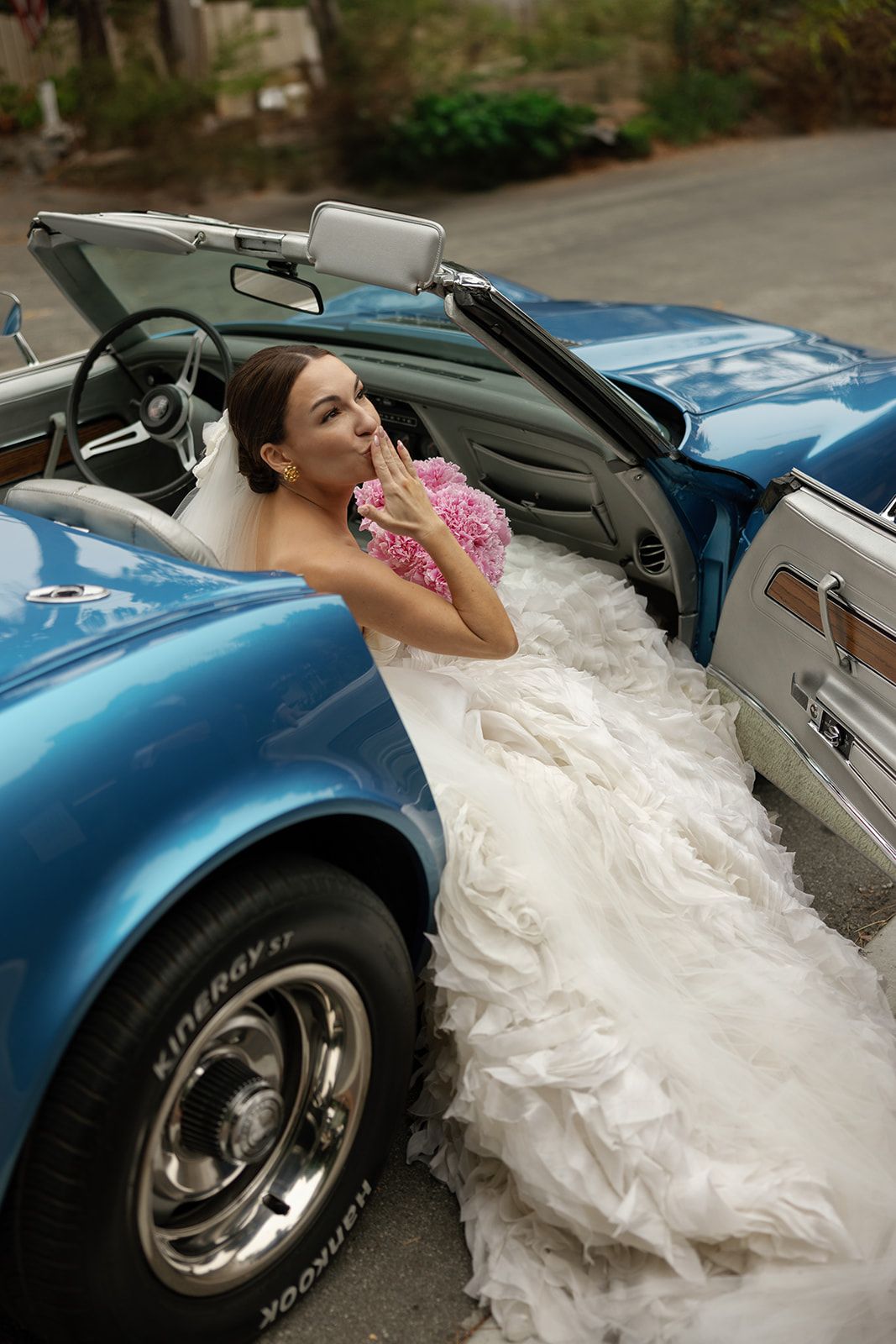 A classic blue convertible car with white wedding dress flowing out of the passenger side on a quiet street.
