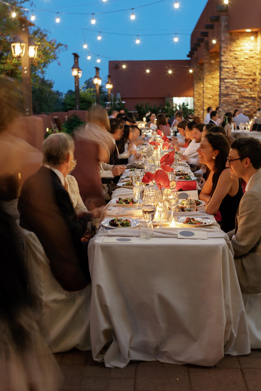 String lights illuminate an outdoor evening dinner party with guests seated at elegant tables with white linens.