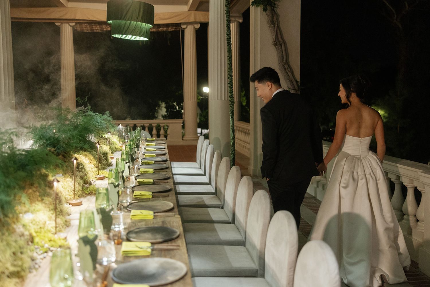Elegant wedding reception dinner table setup with white chairs and green decor, bride and groom admiring the arrangement.
