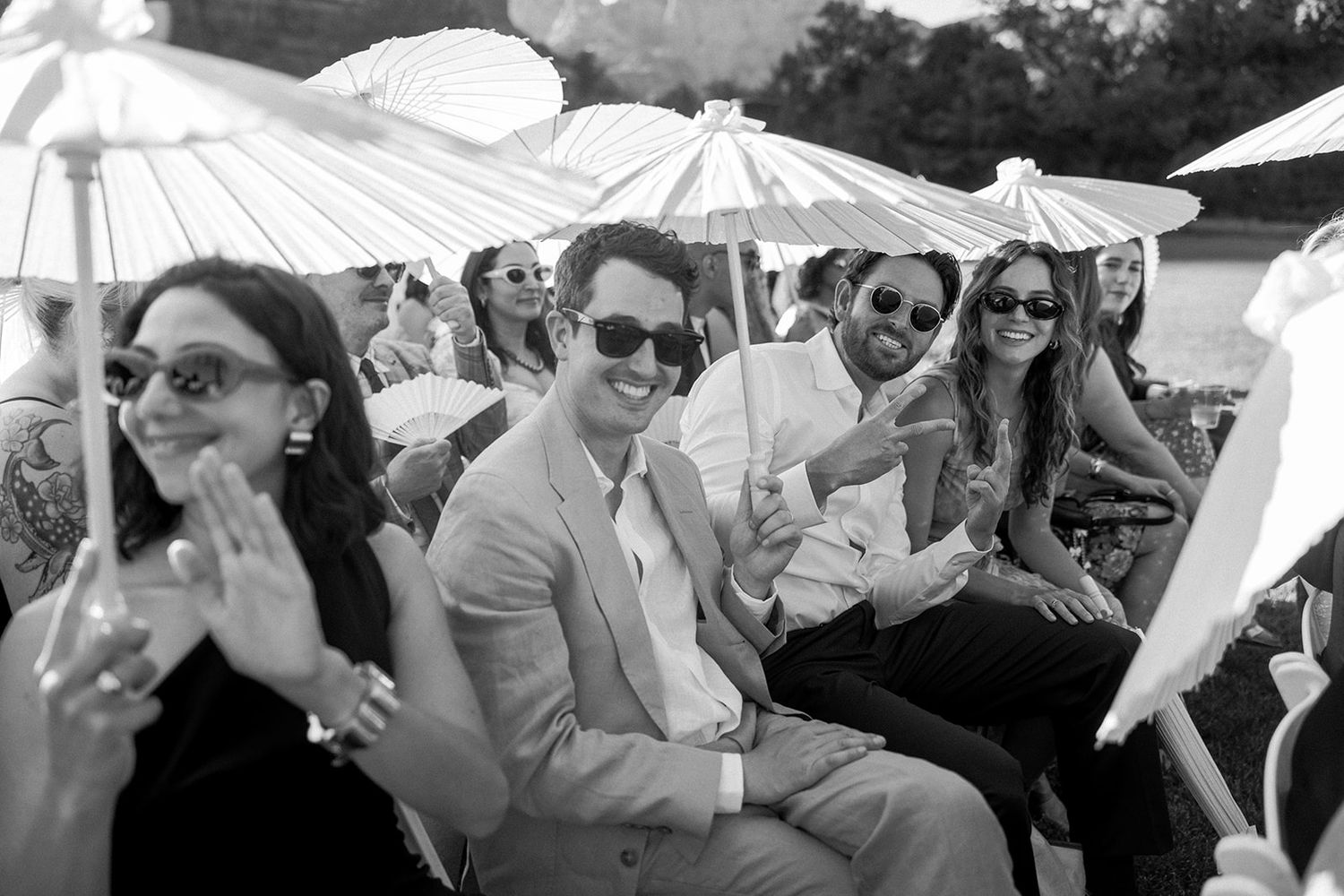 Wedding guests seated outdoors holding white parasols smile and laugh during a sunny celebration.