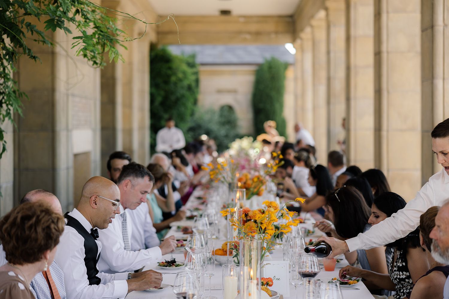 Wedding guests dining at long outdoor table decorated with yellow sunflower centerpieces.