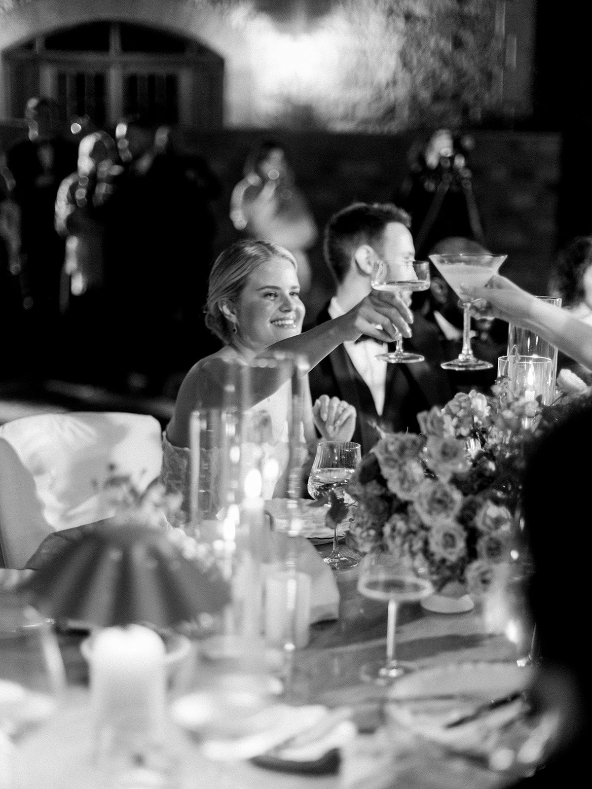Black and white wedding reception toast with smiling bride, groom in tuxedo, candles, and floral centerpieces.