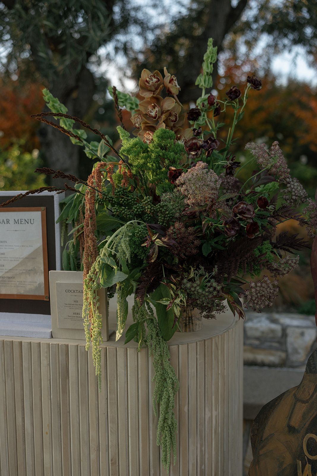 Rustic wooden menu display decorated with cascading greenery and floral arrangements at an outdoor event.