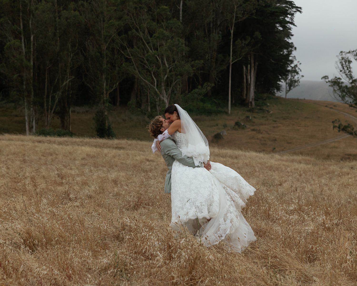 Bride and groom embracing in a golden wheat field with misty forest backdrop on their wedding day.