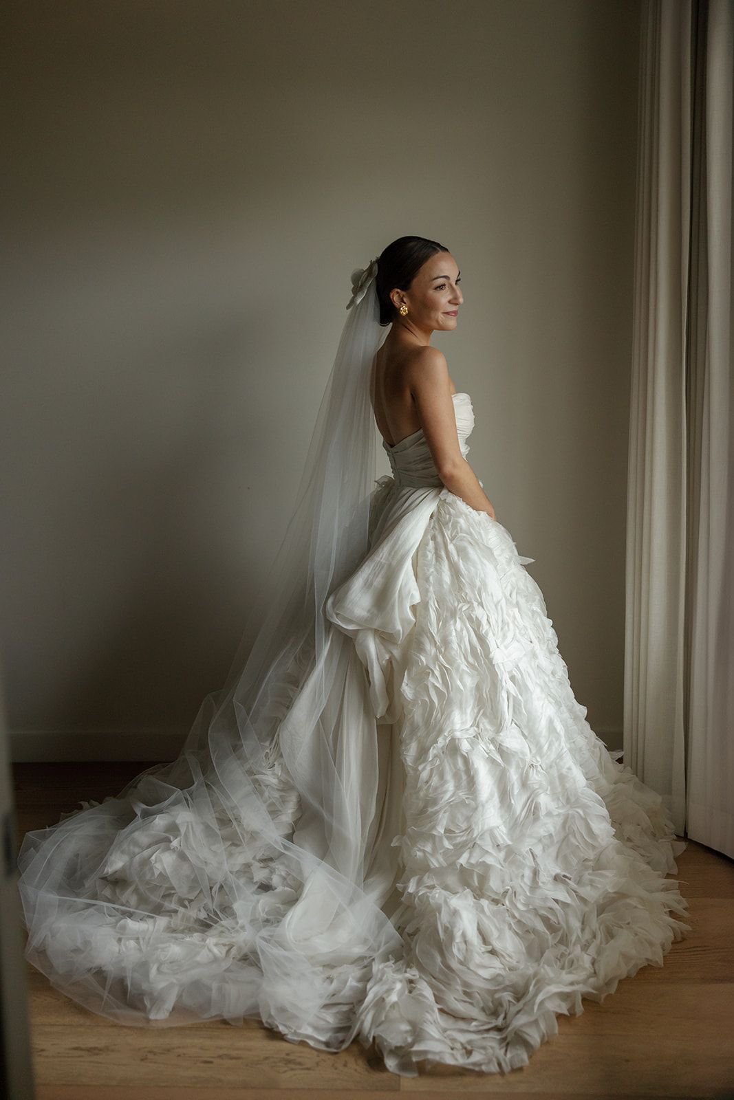 Elegant bridal portraits show a wedding dress's flowing train and ruffles against white walls in indoor natural light.