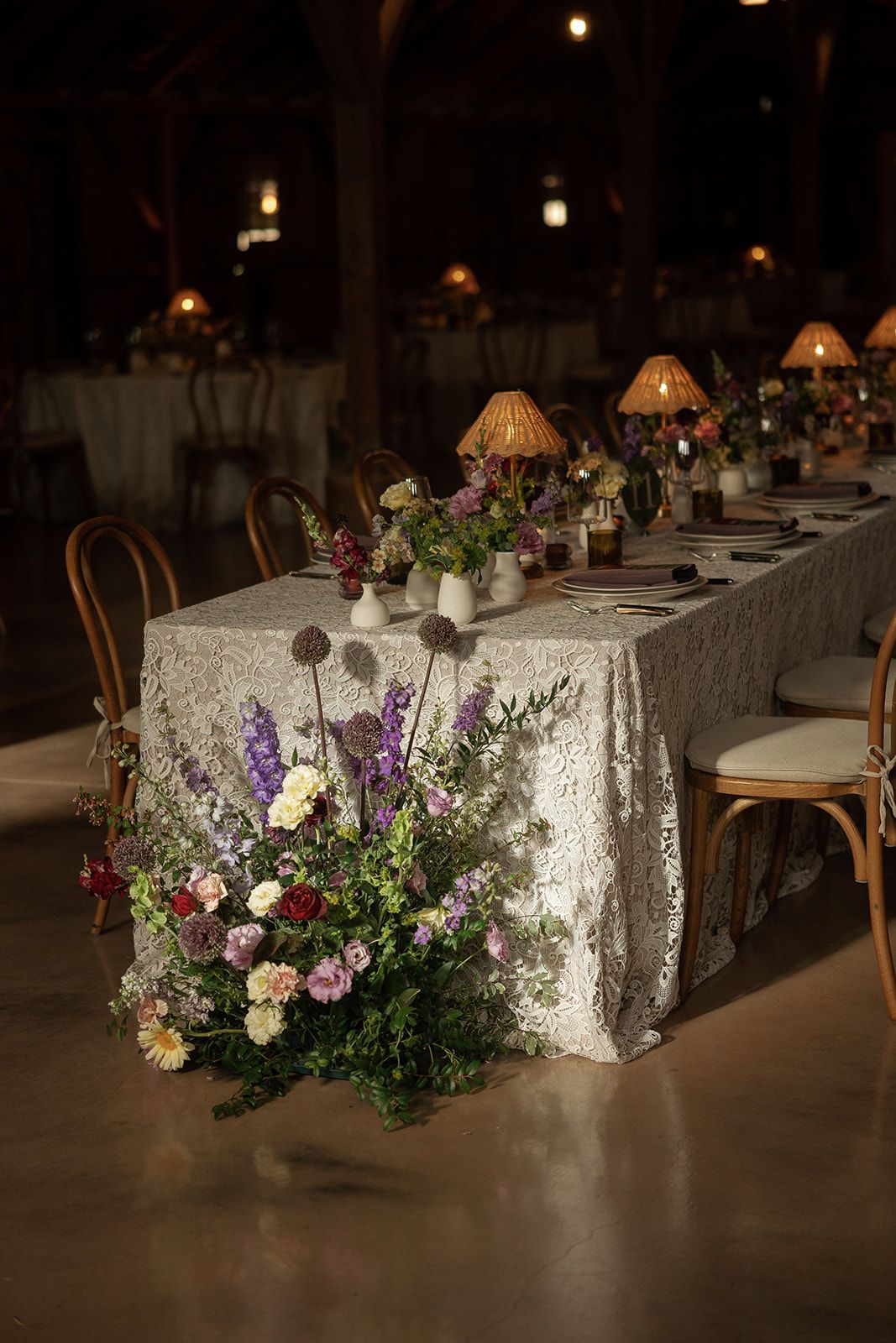 Elegant wedding reception table with lace tablecloth, wildflower arrangements, and warm candlelight in a rustic barn venue.