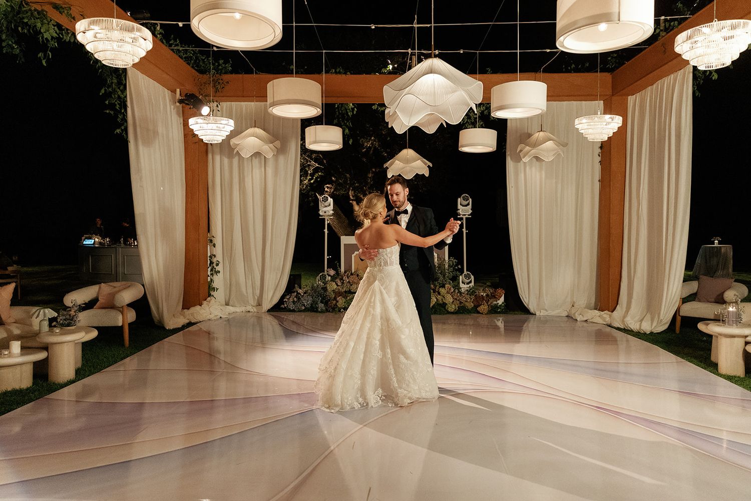 Bride and groom share first dance on white marble floor under elegant chandeliers at night.