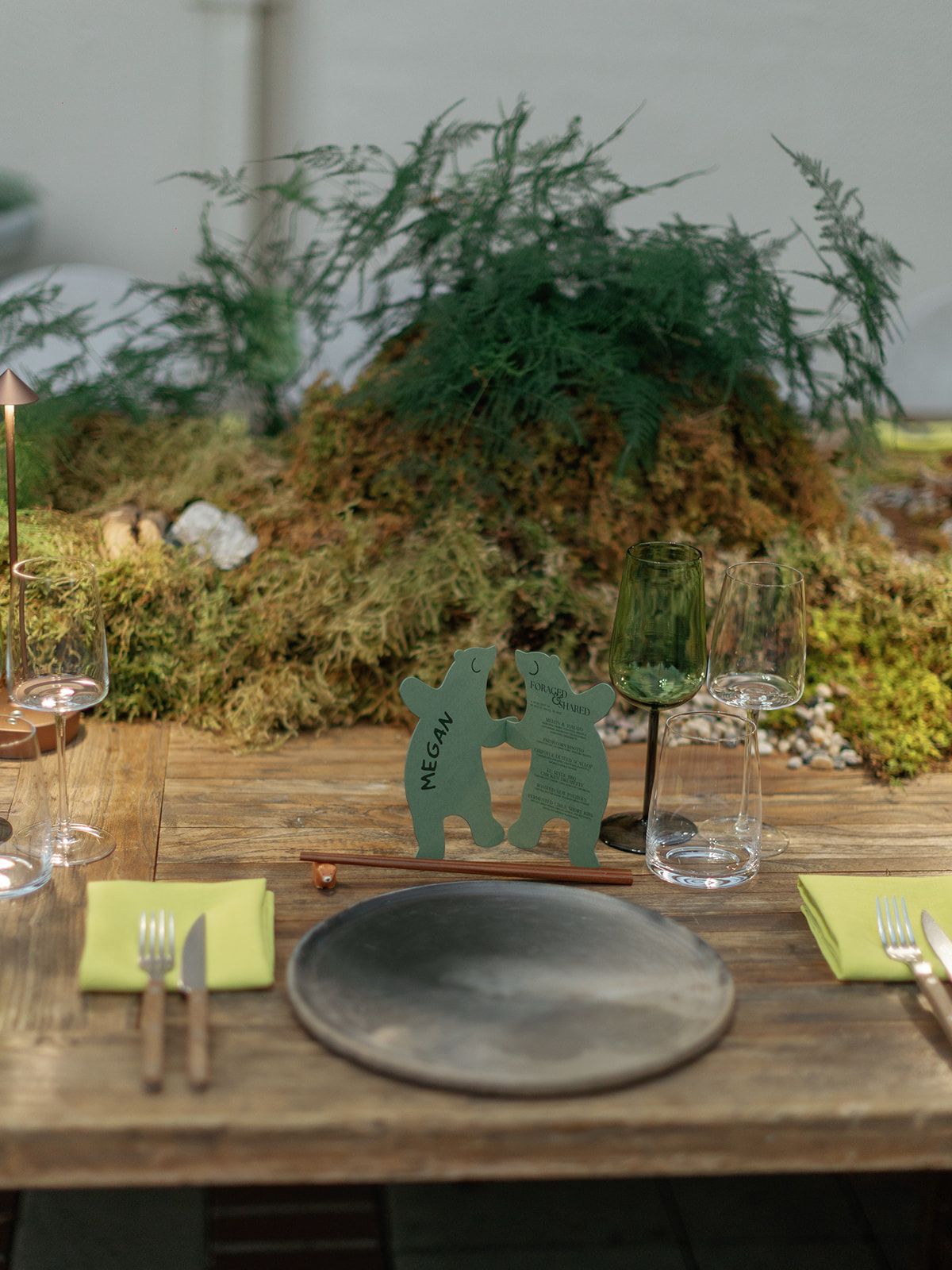 Rustic wooden wedding table setting with green moss centerpiece, dark plate, lime napkins, and a decorative couple silhouette.