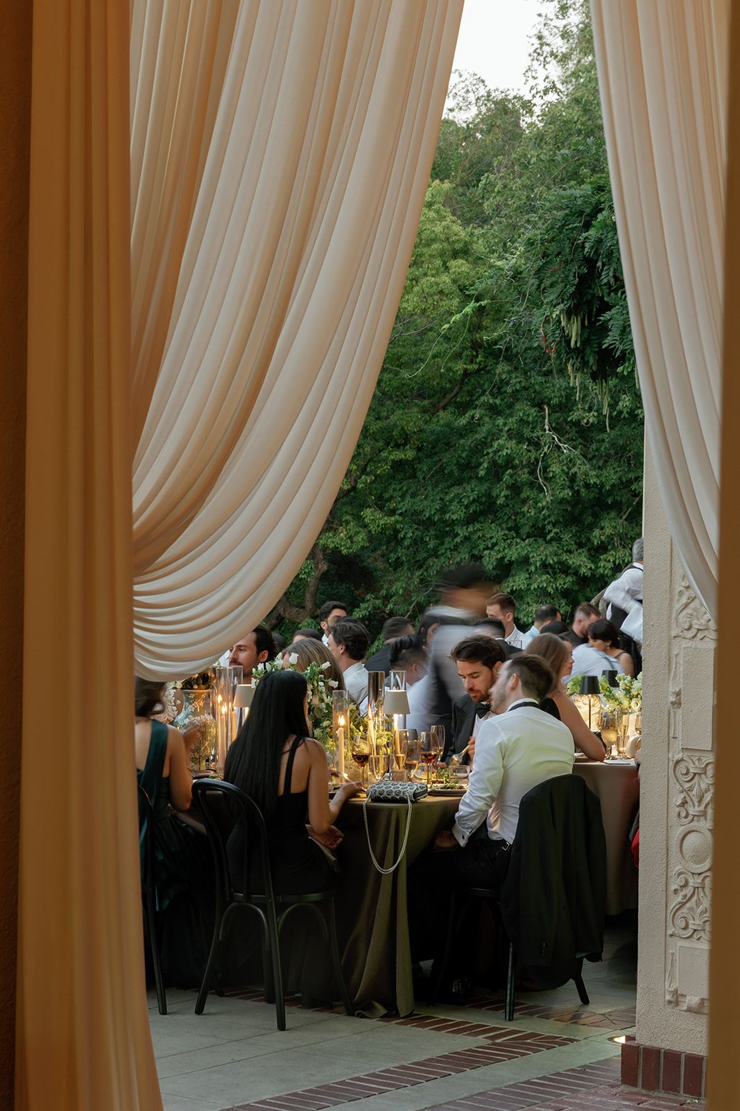 Elegant wedding reception setup with flowing ivory drapes and candlelit tables in an outdoor garden setting at dusk.