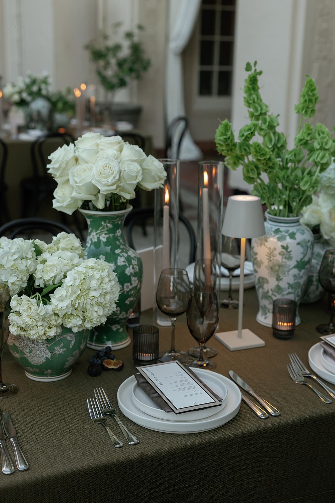 Elegant wedding table setting with dark tablecloth, white roses, greenery and candlelight creates romantic ambiance.