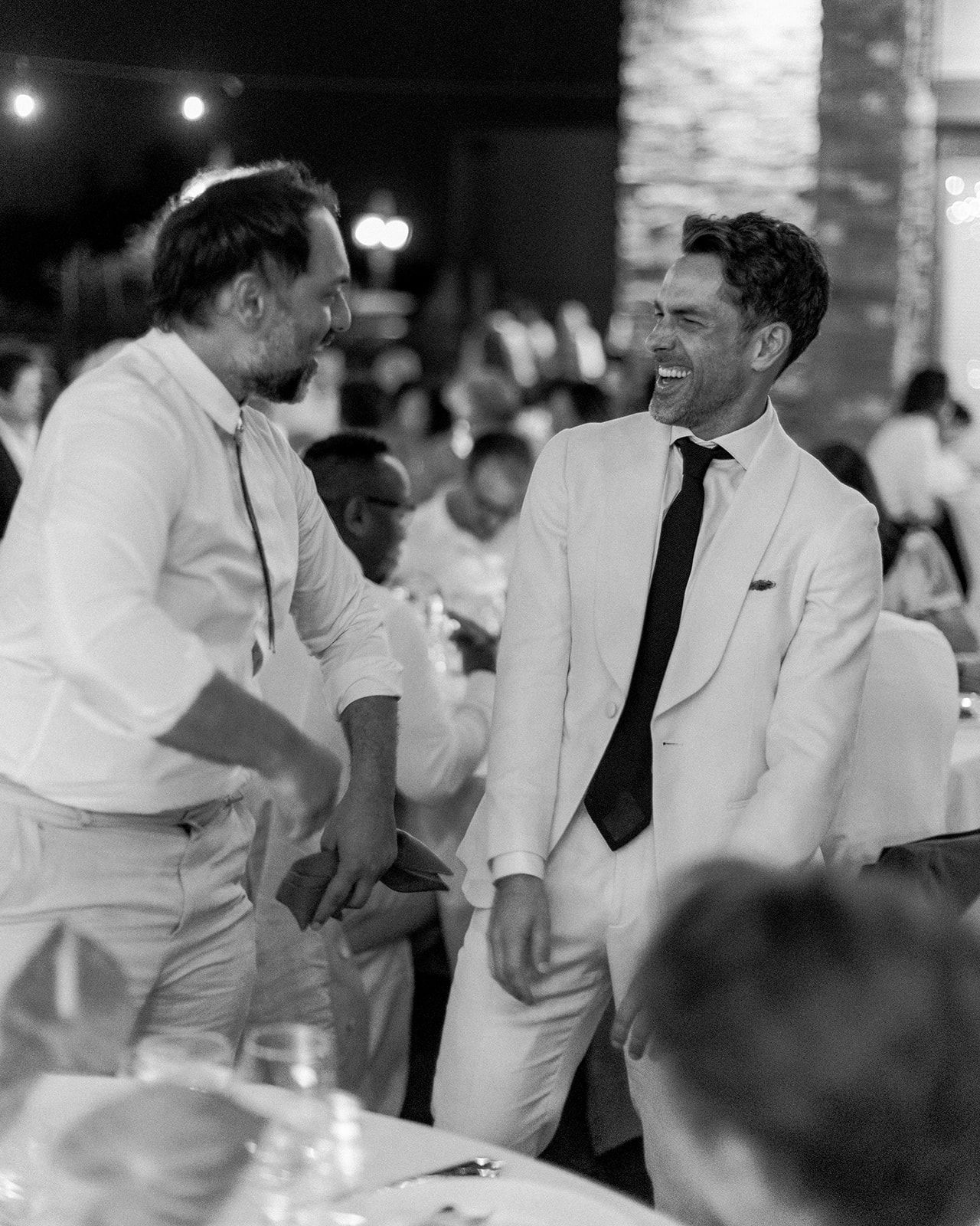 Two men in white suits share a candid moment of laughter at a formal event in black and white photography.