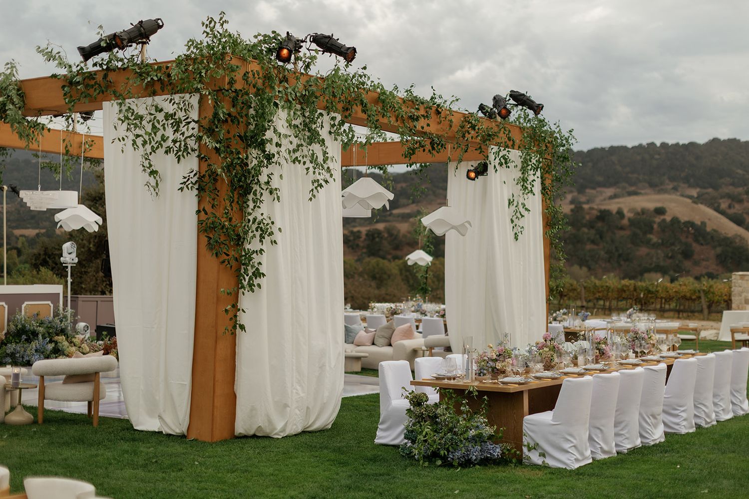 Elegant outdoor wedding reception with wooden pergola, white drapes, and lush greenery on a hillside.