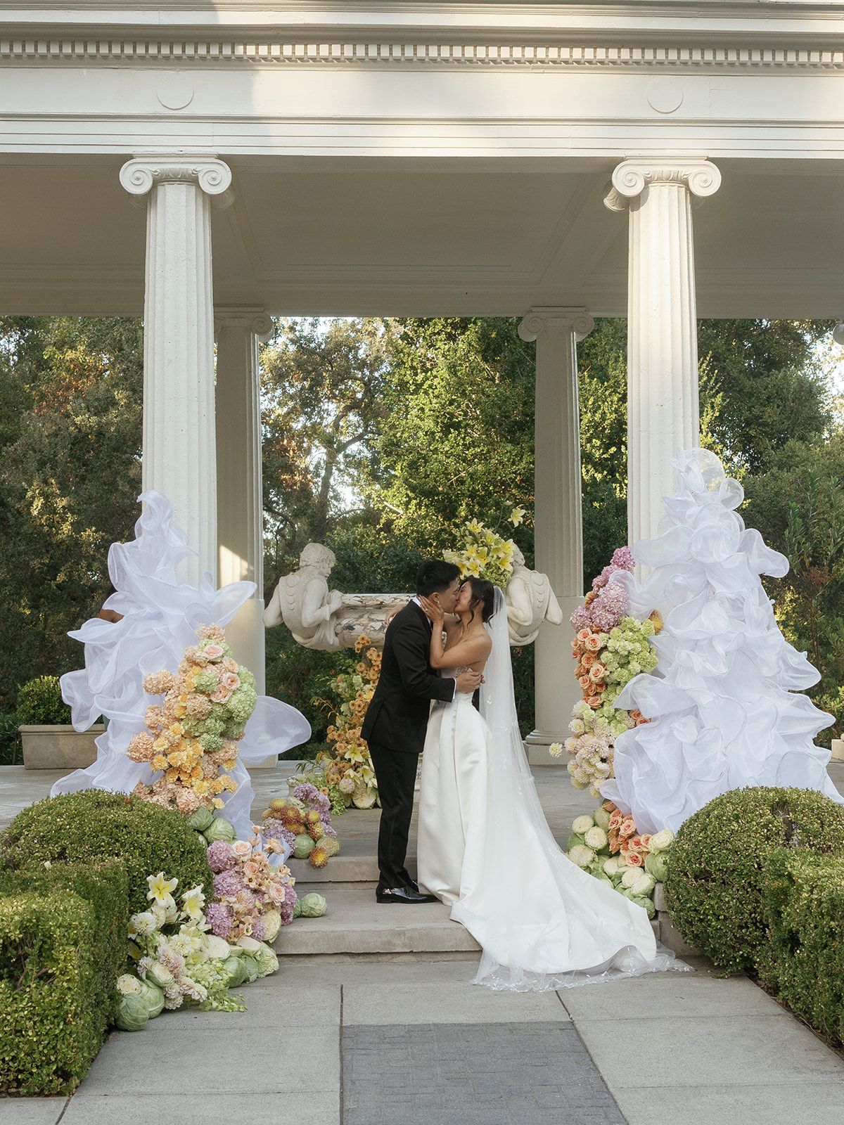 Bride and groom kissing at elegant outdoor wedding ceremony with floral arches, white columns, and lush garden greenery.