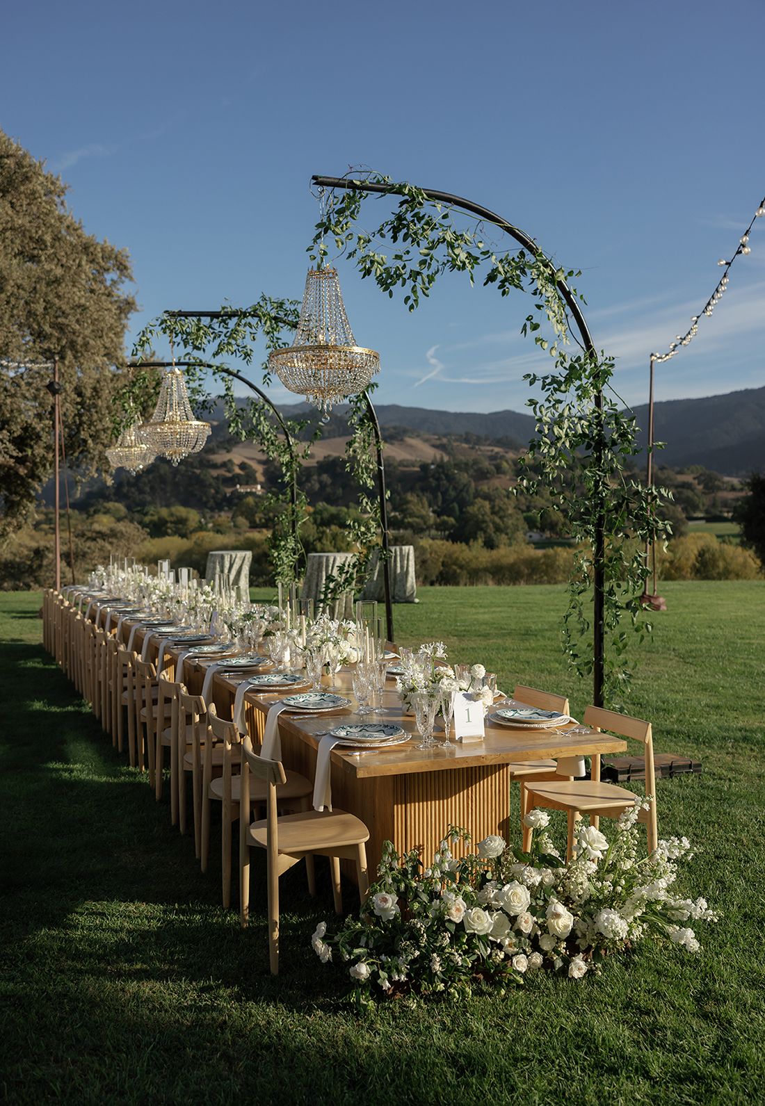 Long rustic wooden dining tables set outdoors with elegant white floral arrangements and arched greenery against mountain views.