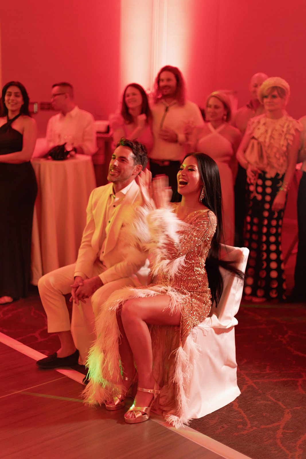 A couple in glamorous outfits sitting together at a party bathed in red lighting while others look on from behind.