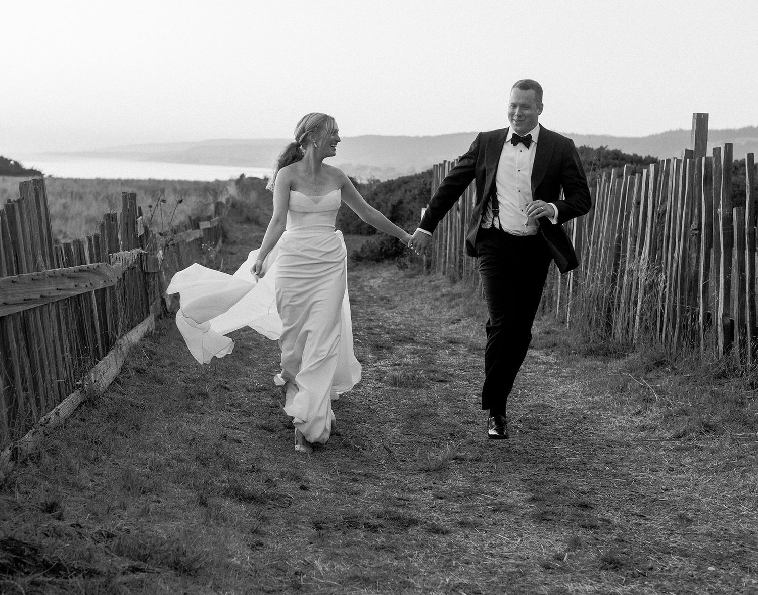 Elegant couple runs playfully along rustic fence path with bride's dress flowing behind them.