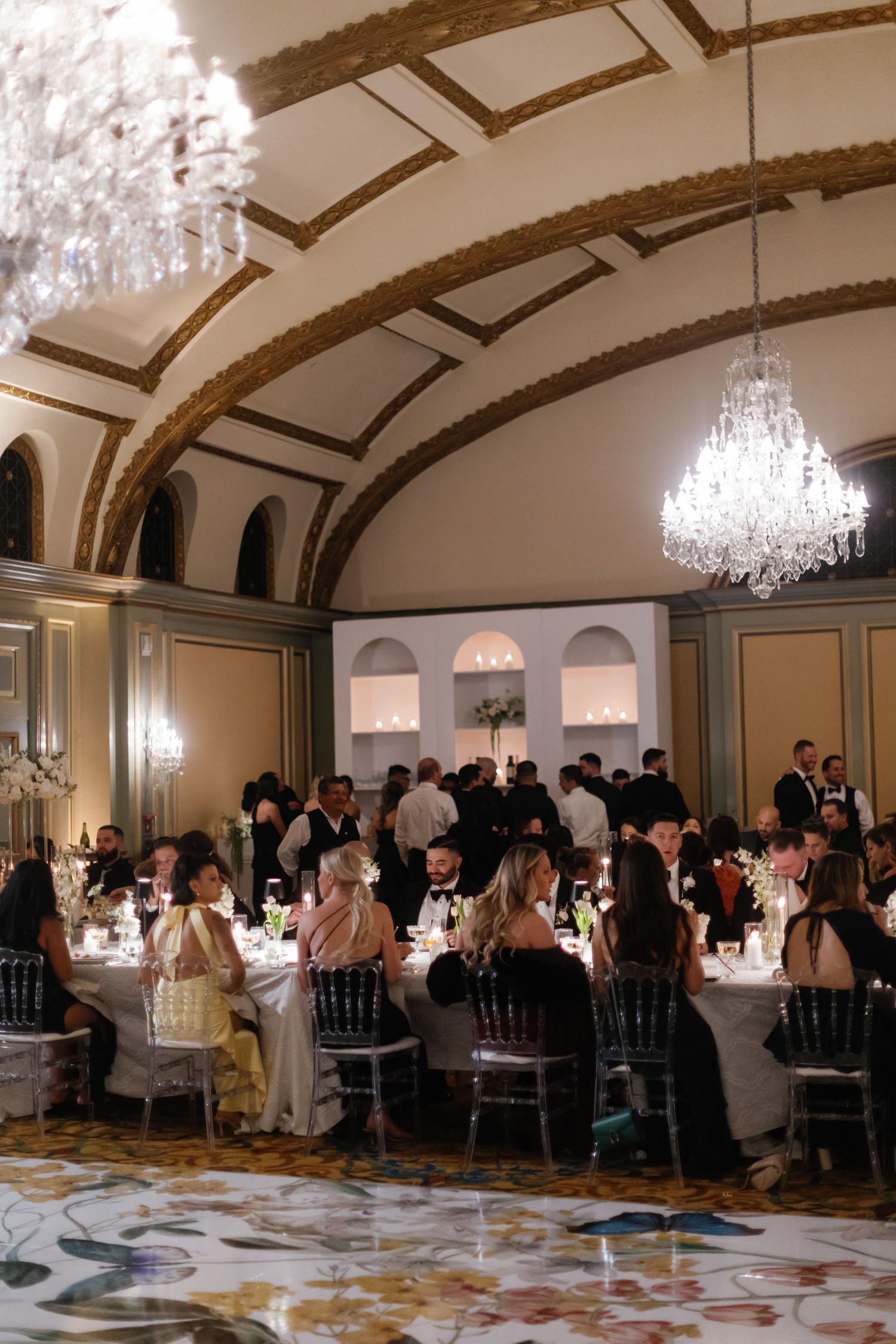 Elegant wedding reception in a grand ballroom with arched ceilings, crystal chandeliers, and guests dining at round tables.