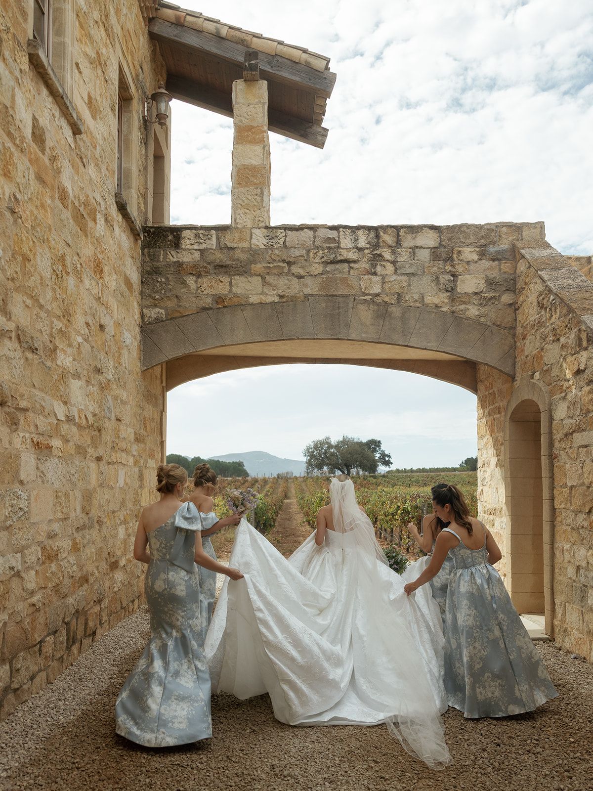 Bride in white gown and veil with bridesmaids in blue dresses under stone arch at vineyard wedding venue.