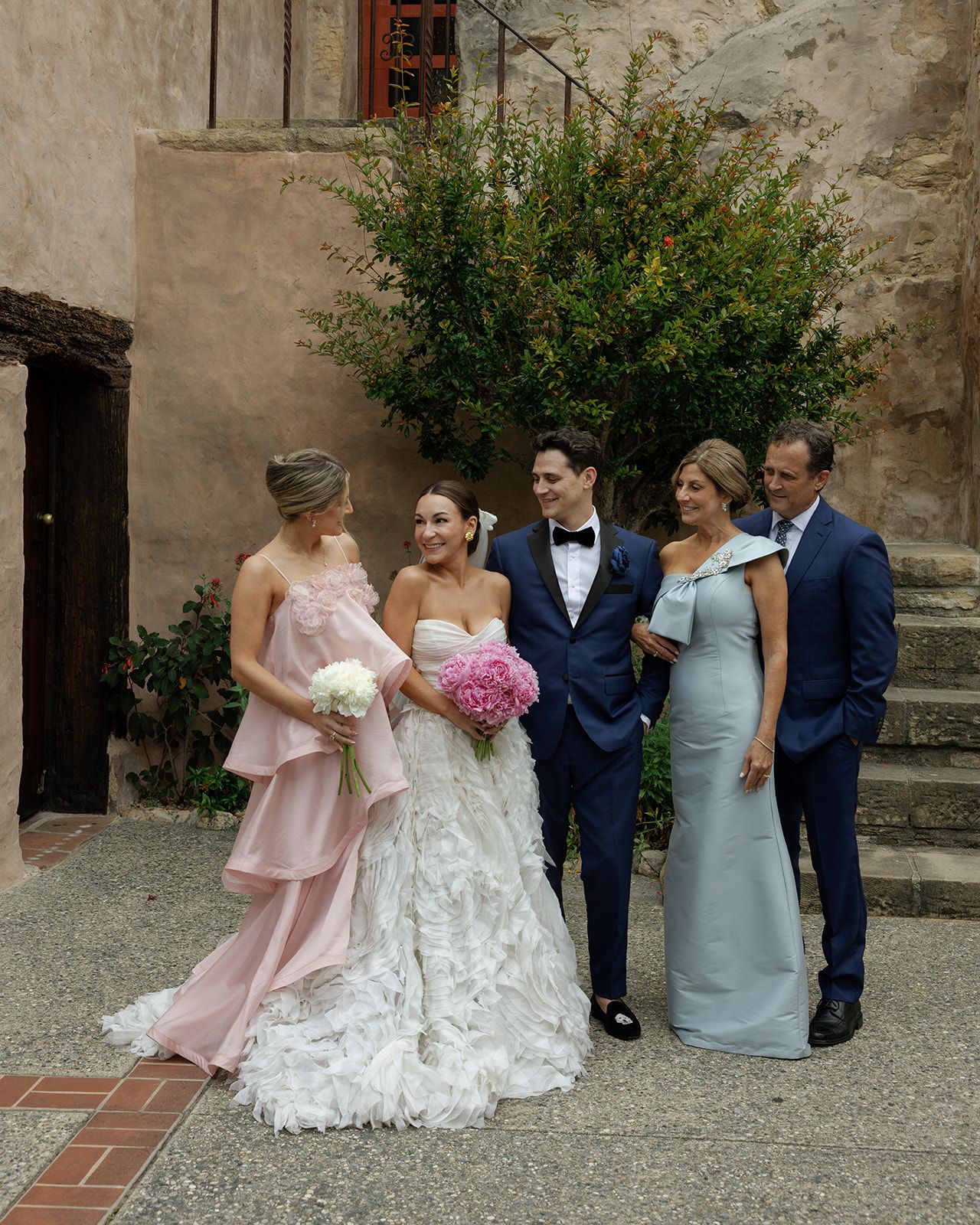 Wedding party in formal attire laughing together against an old stone wall in a historic courtyard setting.