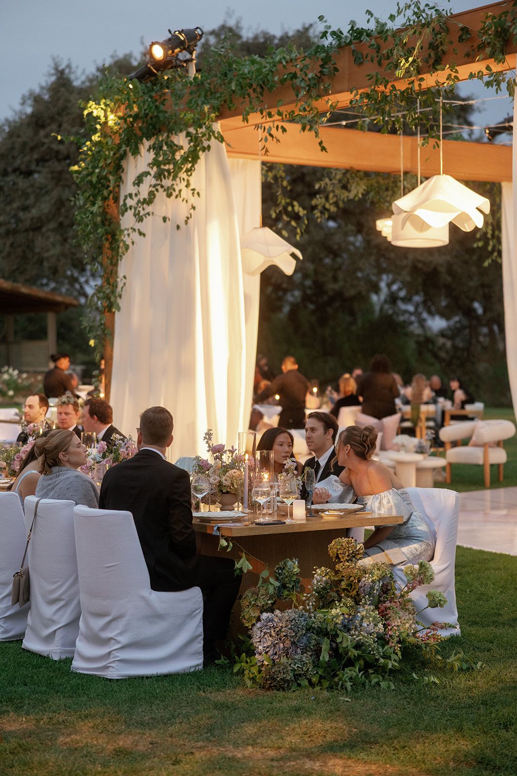 Elegant outdoor wedding reception at dusk with floral arrangements, white draping, and guests seated at a decorated table under a pergola.