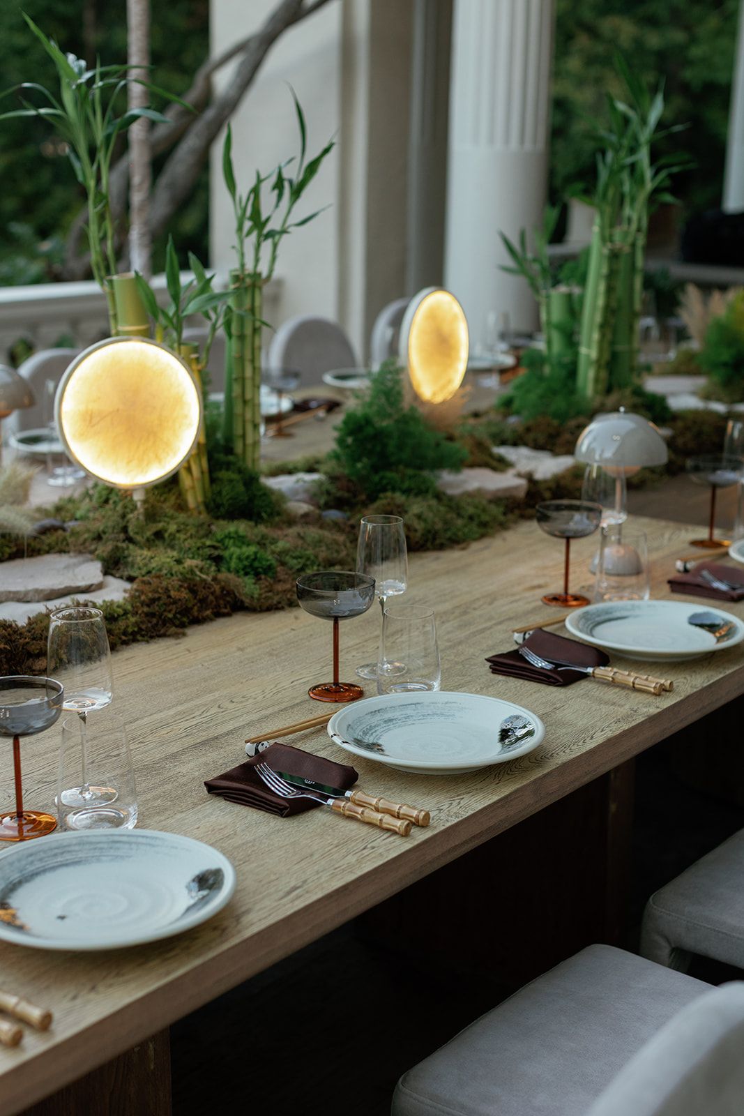 Elegant dining table with glowing circular lamps, bamboo plants, marble plates, and crystal glasses on a wooden surface.