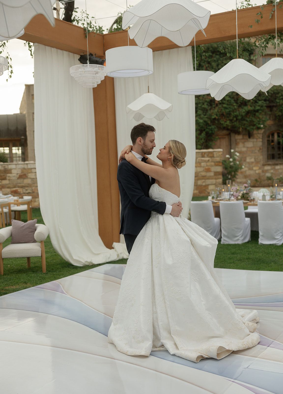 Bride and groom share first dance outdoors at elegant wedding reception with white draping and pendant lights.